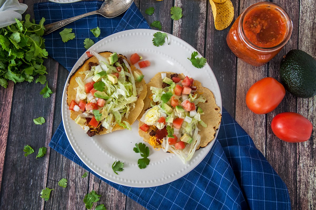 Holding one of two tacos over a plate. The plate is on a table that contain a bunch of taco fixings.