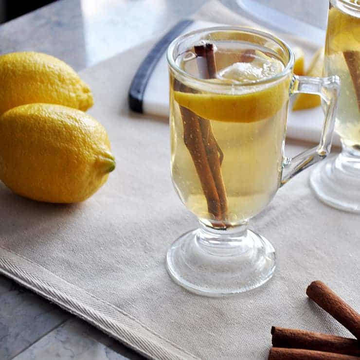 backlit picture of a hot toddy on tablecloth with two lemons and some cinnamon sticks.