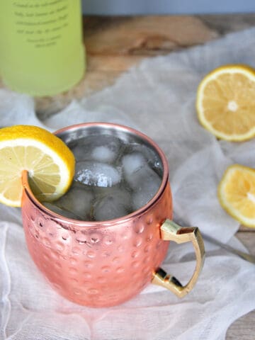 Limoncello and Lavender Moscow Mule on a wood table with a bottle of limoncello in the background. There is a sliced lemon on the table and a stirring spoon.