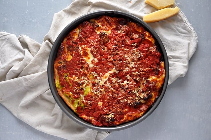 A Chicago Style Pizza in a pan and on a white kitchen towel. Some parmesan cheese is sitting on the side