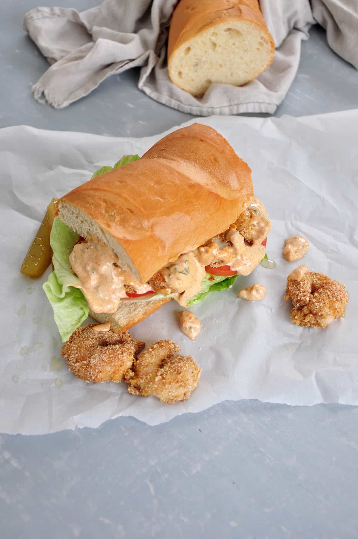 Finished po' boy sandwich with remoulade sauce on it. French bread in background wrapped in cloth.