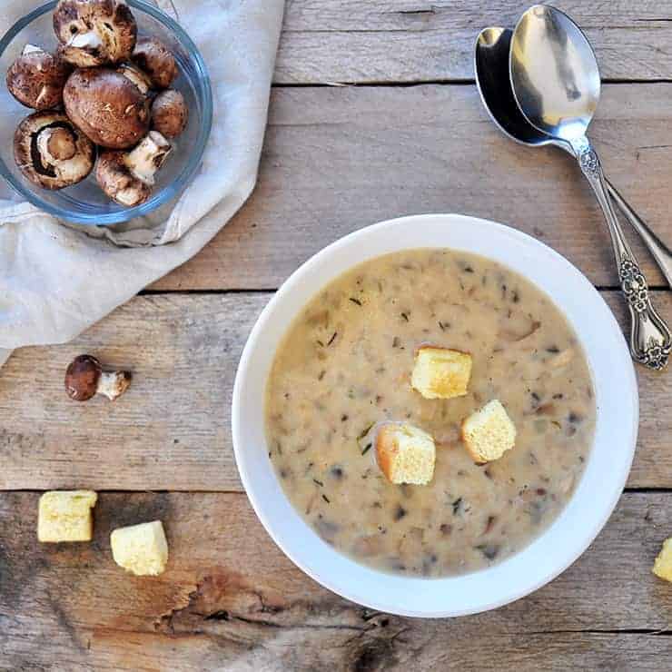 Overhead shot of cream of mushroom soup with croutons on top and mushrooms on the side. Two spoons on the side and all of it on old wood.