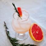 Rosemary infused Salty Dog on a mat. Half a grapefruit close by and a sprig of rosemary on the side