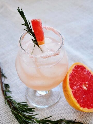 Rosemary infused Salty Dog on a mat. Half a grapefruit close by and a sprig of rosemary on the side