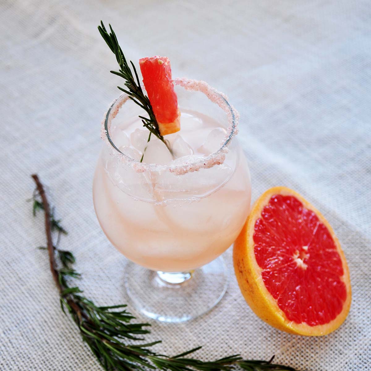 Rosemary infused Salty Dog on a mat. Half a grapefruit close by and a sprig of rosemary on the side.