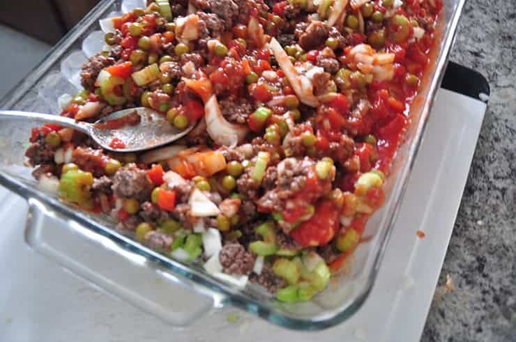 mixed hotdish that still needs to be baked. 9 by 13 dish on cutting board with a spoon in the dish mixing.