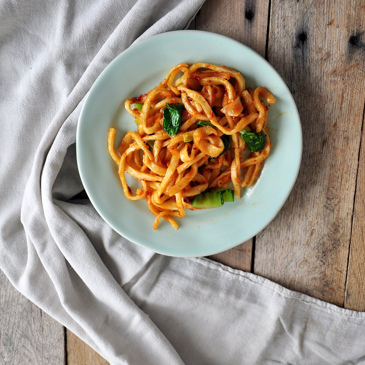 chow mein noodles with bok choy on a turquoise plate. This is on a wood background with a white cloth.