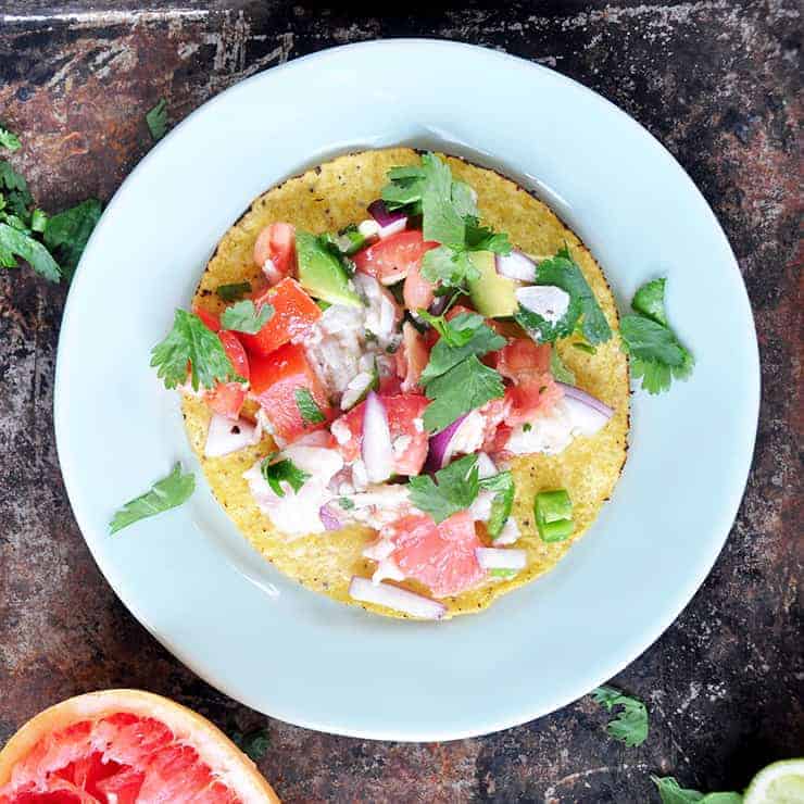 Grapefruit ceviche on a tostada that is also on a white plate with a grapefruit in the background and cilantro sprinkled on the plate