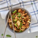 looking down on a bowl of crispy chicken chow mein in a grey bowl. There are chop sticks on top of the bowl, a plaid napkin is under the bowl. Green onions are on the table next to the bow.