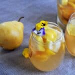 Glass of our best white sangria recipe with some viola flowers garnishing it with a pear and pitcher in the background