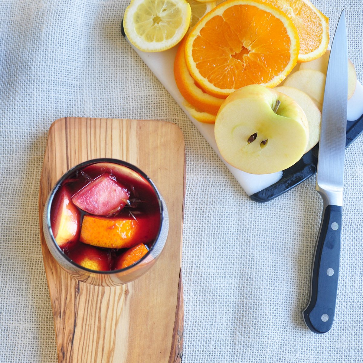 Glass with sangria and fruit in it on a piece of wood. There is a cutting board with oranges, lemons and an apple on it.