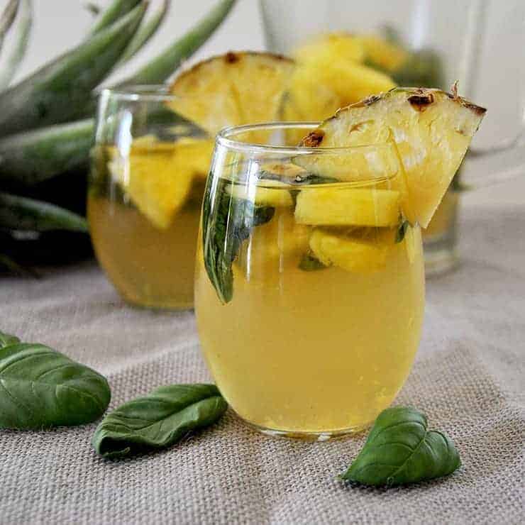 Two glasses of basil pineapple moscato sangria with some basil laying on a mat and pineapple head in the background