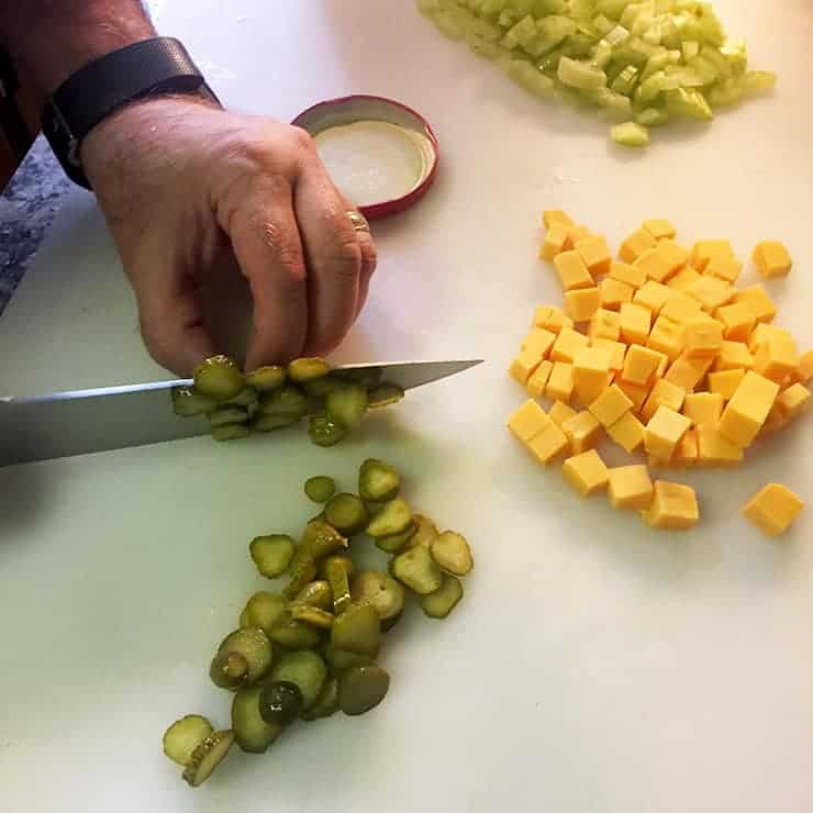 cutting board with ben myhre chopping pickles. There is diced cheese and chopped celery on the same chopping board.