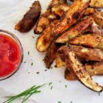 A plate of grilled jo jos with a small bowl of ketchup and some chives. All of this is on some parchment paper.