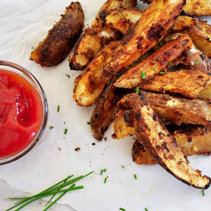 A plate of grilled jo jos with a small bowl of ketchup and some chives. All of this is on some parchment paper.