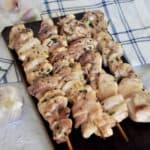 Chicken Shish Kabob on a wood plank with garlic, rosemary, and thyme in the background