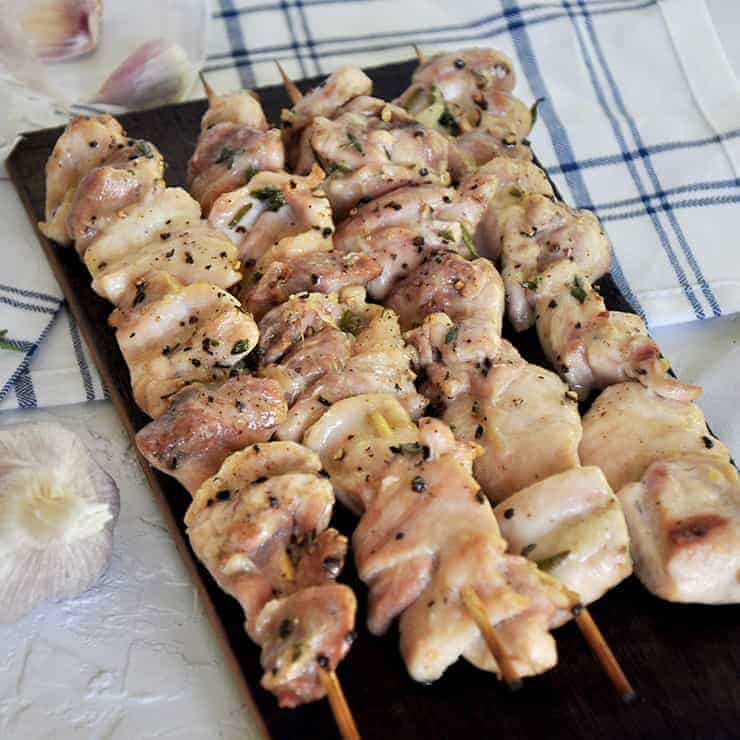 Chicken Shish Kabob on a wood plank with garlic, rosemary, and thyme in the background.