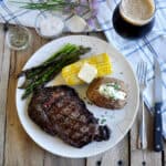 Overhead view of Ribeye Steak on a plate with a corn cob, baked potato, asparagus, and a Russian Imperial Stout