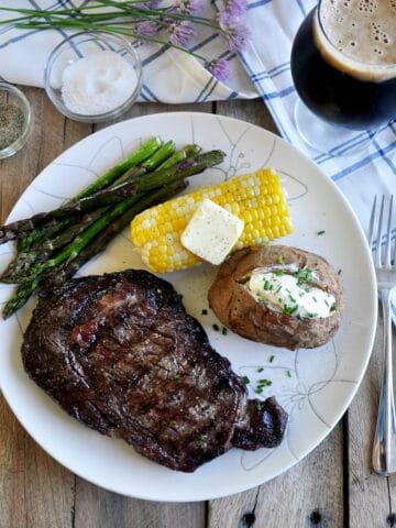 Overhead view of Ribeye Steak on a plate with a corn cob, baked potato, asparagus, and a Russian Imperial Stout