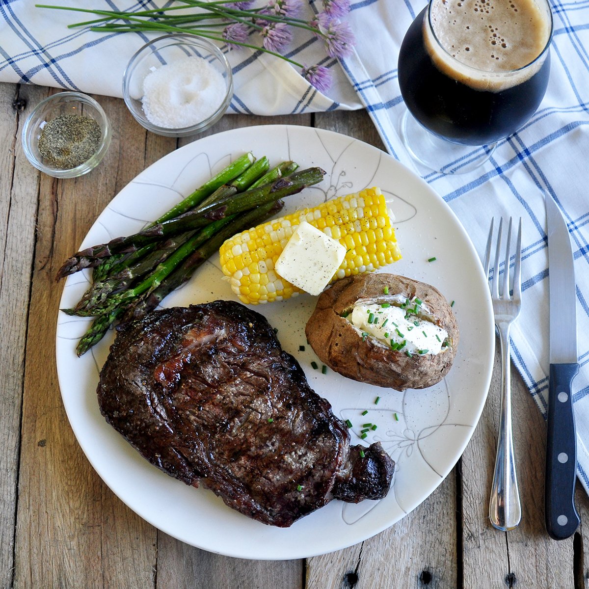 Overhead view of Ribeye Steak on a plate with a corn cob, baked potato, asparagus, and a Russian Imperial Stout.