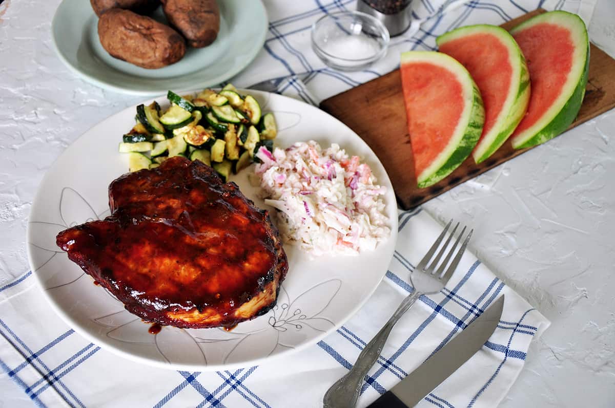 completed pork chops on a plate with grilled zucchini, coleslaw, and sliced watermelon in the background.