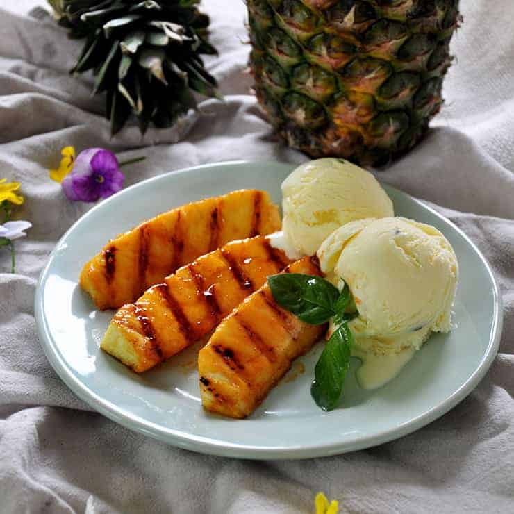 Grilled pineapple with a side of basil ice cream on a plate with pineapple in the background and some decorative flowers.