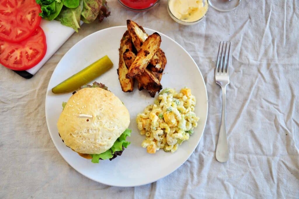 overhead burger shot on a white plate, macaroni salad, jojo potatoes, a pickle, and tomatoes