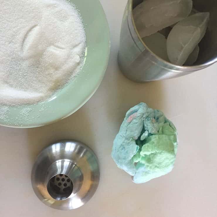 Cotton Candy Margarita ingredients on a table, with a cocktail shaker and a plate that has sugar for rimming