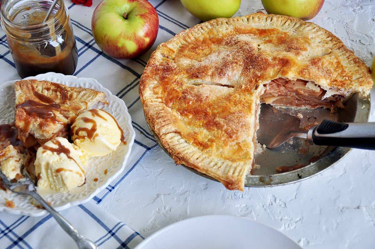 Baked apple pie with two pieces removed and placed on a plate. There is a serving utensil in the pie pan. Several Braeburn apples in the background