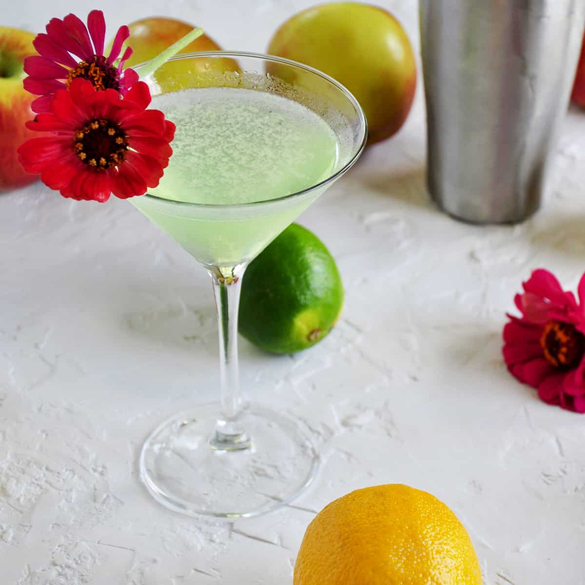 Appletini with lemons, limes, apples and flowers in the background.