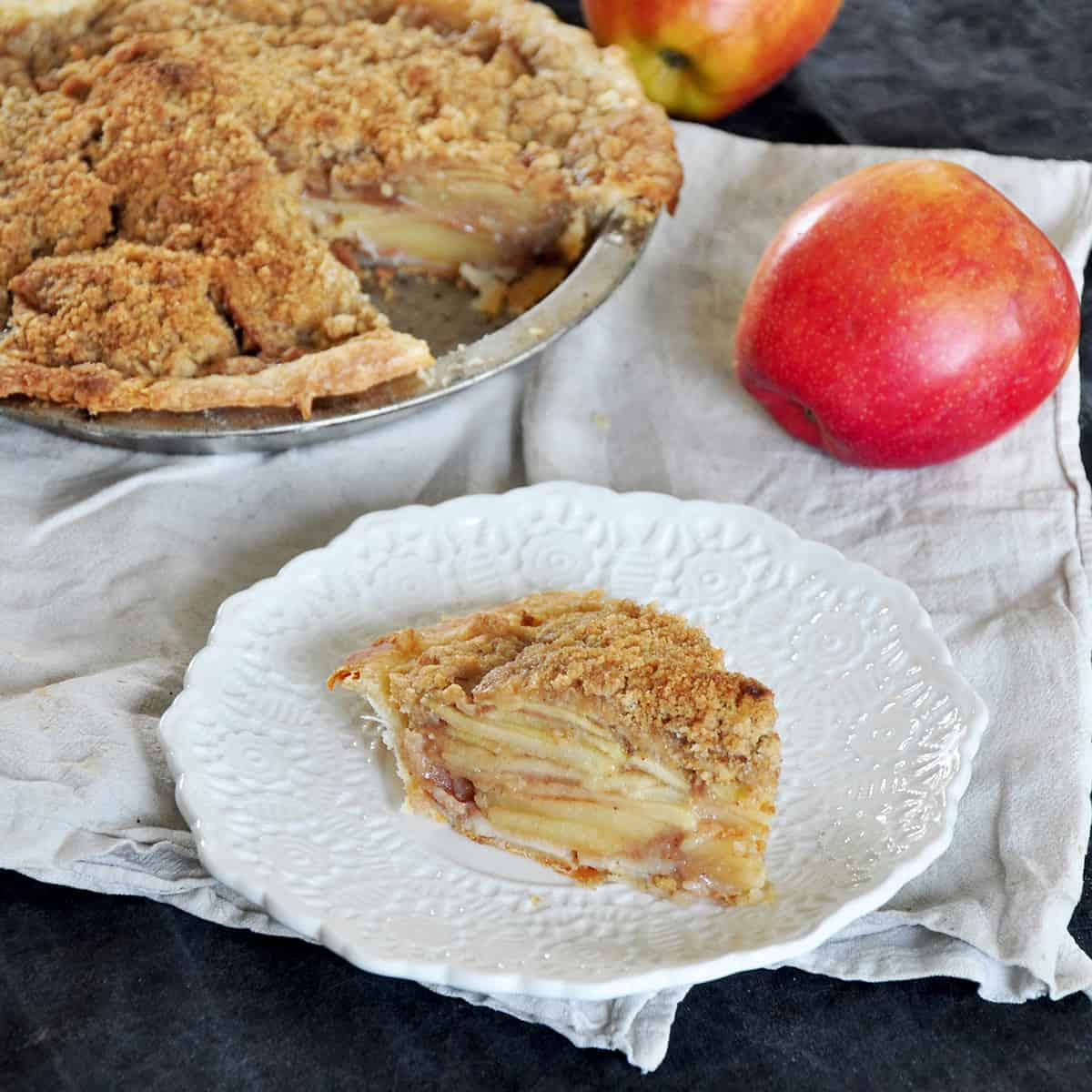 A single slice of a dutch apple pie with the rest showing in the background. There is a single Braeburn apple in the back and it is all on a white cloth.