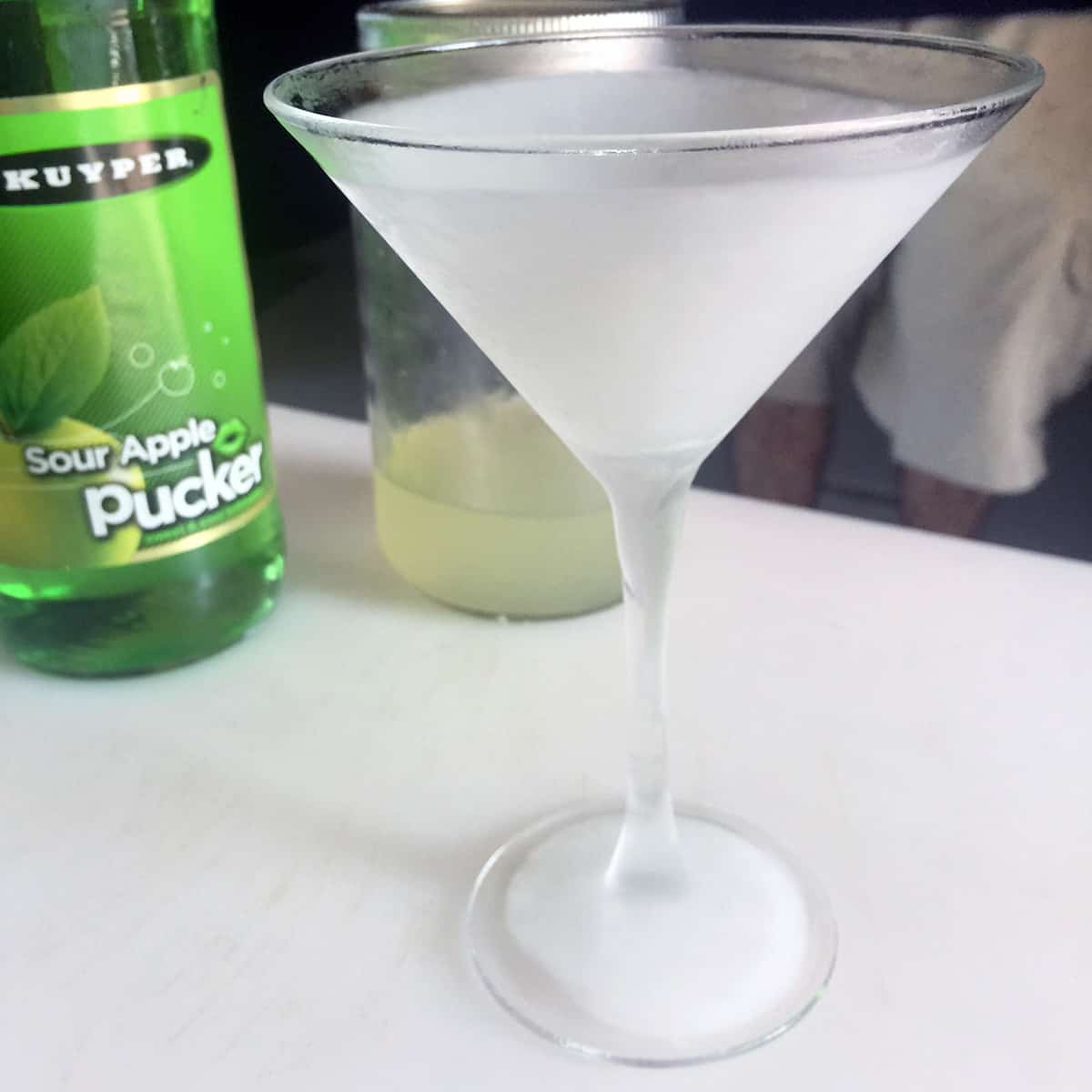 A frozen martini glass with a bottle of apple pucker in the background.