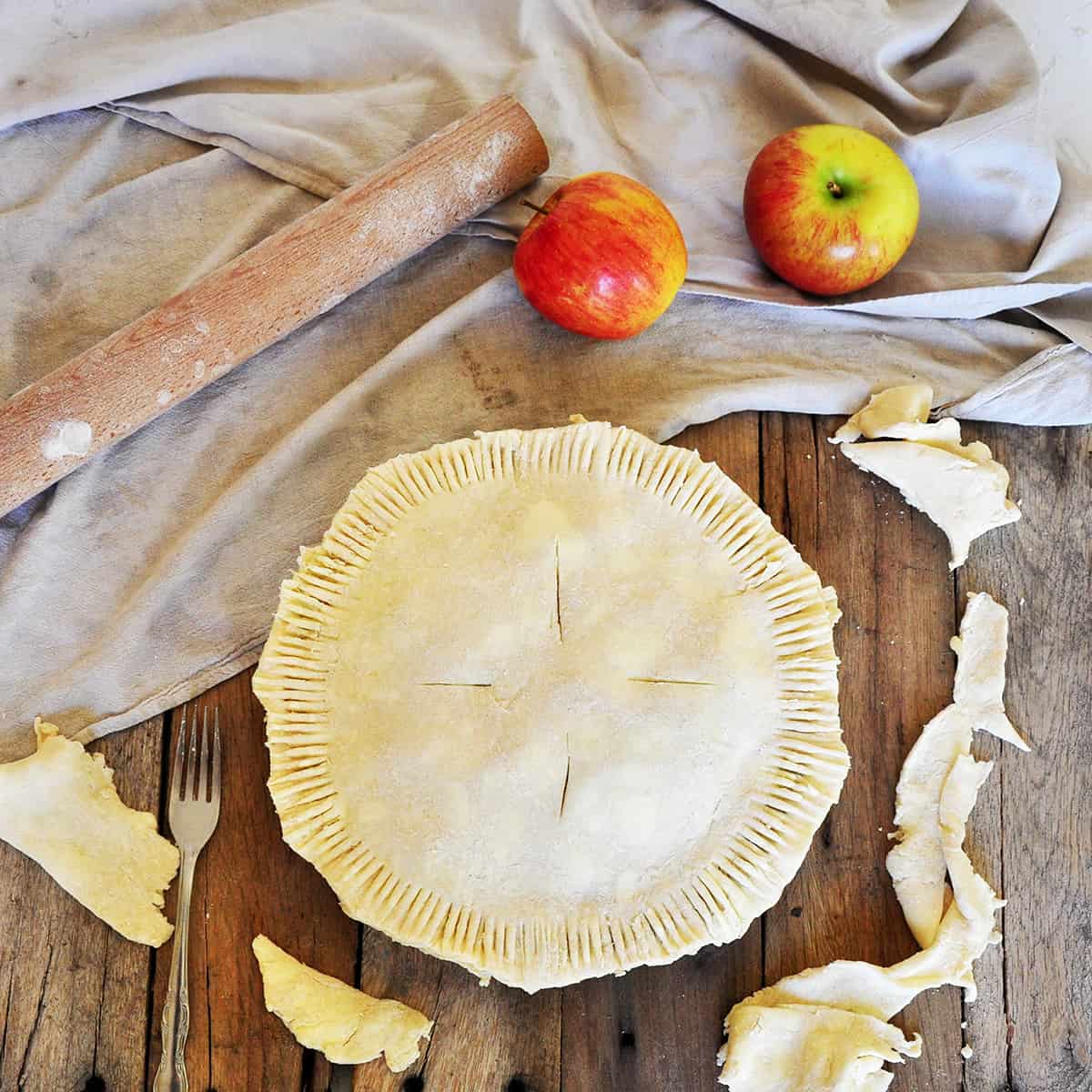 A Raw Pie Crust with apples in the pie. It is on a picnic table with two Braeburn apples, a rolling pin, and is laying on a cloth.