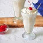 White Russian Milkshake on a white table. There is a second adult cocktail on a wood cutting board right next to a bowl of maraschino cherries.