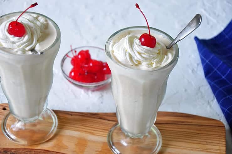 Two shakes on a wood cutting board. They are topped with whipped cream.