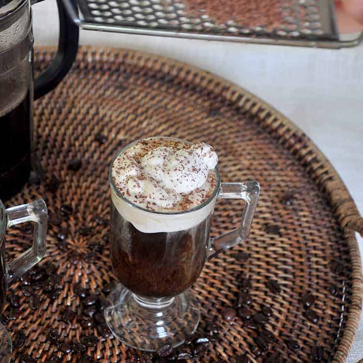 Single Glass of our Russian Coffee recipe on a wicker platter that has coffee beans strewn throughout. I am grating some cocoa on top of the drink for flavor and garnish.