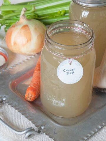 Homemade Chicken Stock in quart jars. There are onions, celery, carrots, and garlic spread around the chicken stock.