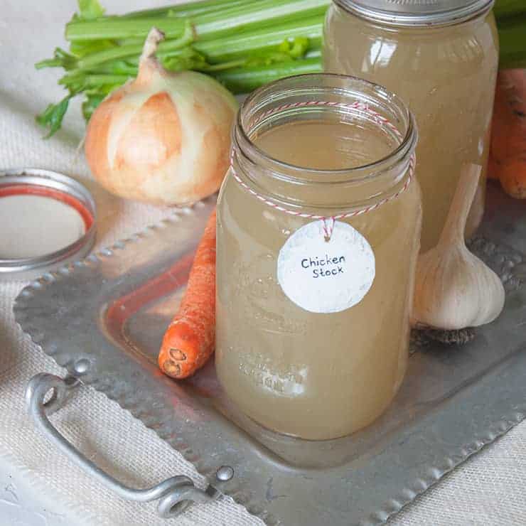 Homemade Chicken Stock in quart jars. There are onions, celery, carrots, and garlic spread around the chicken stock.
