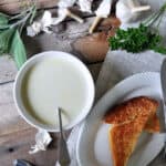 Garlic Soup in a white bowl on a picnic table. On the side, there is a serving of cheesy toast and several fresh herbs for display.