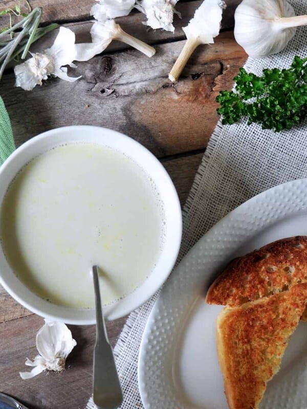 Garlic Soup in a white bowl on a picnic table. On the side, there is a serving of cheesy toast and several fresh herbs for display.