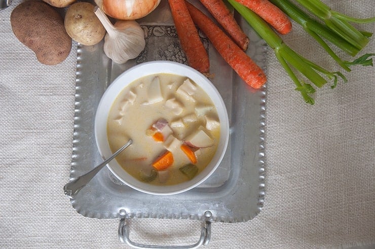 Bowl of Knephla Soup on a silver trey with carrots, potatoes, and celery in the background