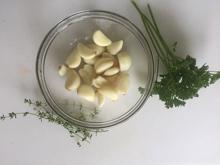 Peeled Garlic in a glass bowl. These cloves of garlic were briefly blanched and then peeled. On the side, there is some thyme and fresh parsley.