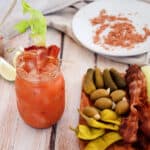 Bacon Bloody Mary with a tray of pickles and olives. In the background is the bacon salt that is used on the drink