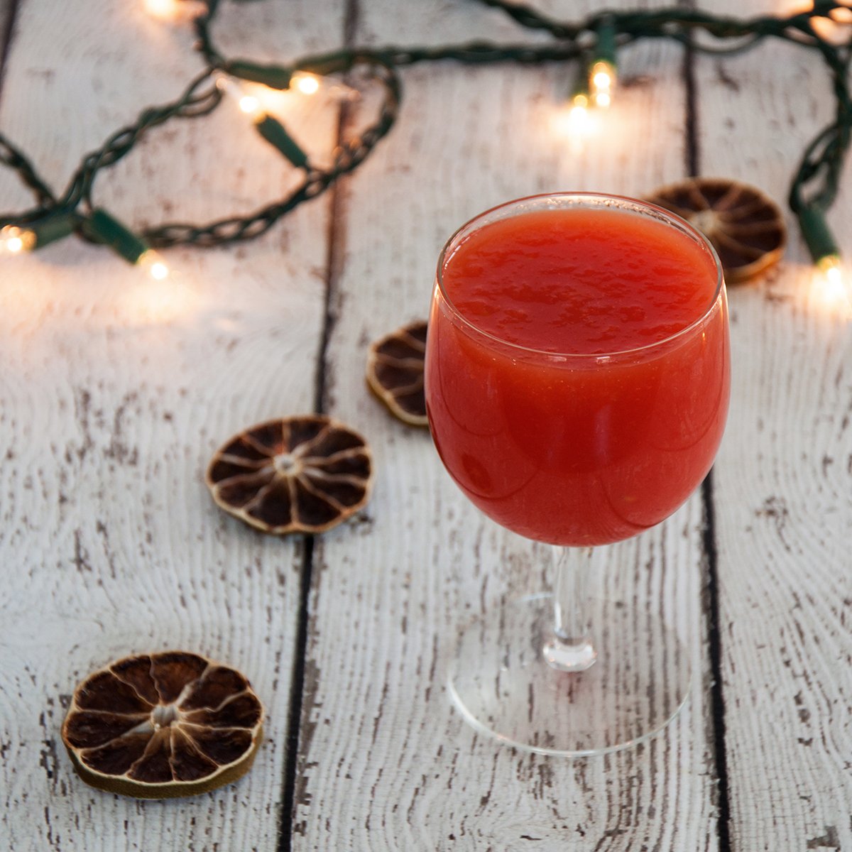 A glass of tomato juice on a wood table with dried limes and holiday lights in the background