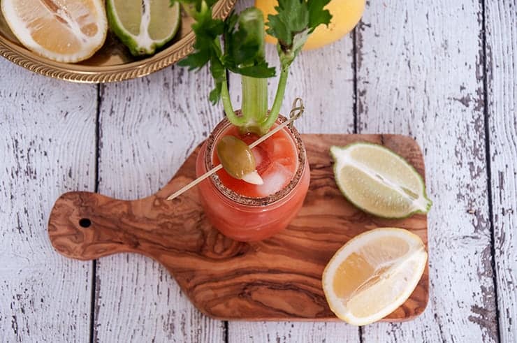 Overhead image of a Bloody Caesar cocktail with lemon, lime, olive, and celery in the background