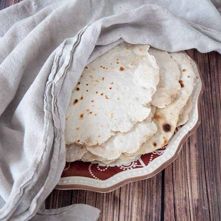 Homemade flour tortillas on a white and red plate with a white towel covering