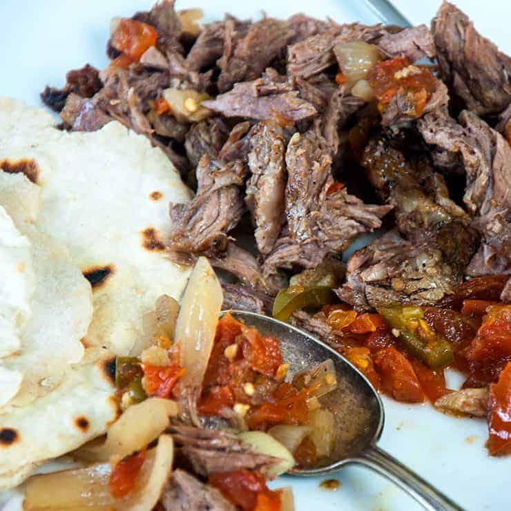 Shredded beef on a cutting board with some of the fixings and flour tortilla on the side