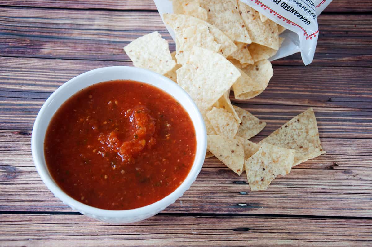 A bowl of salsa with some corn tortilla chips right next to it on a wood table.