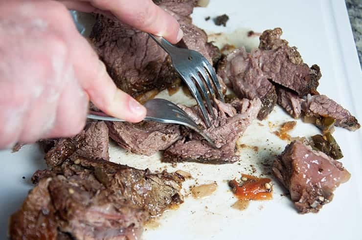 Two forks being held and the prongs are being used to pull the beef apart on a cutting board.