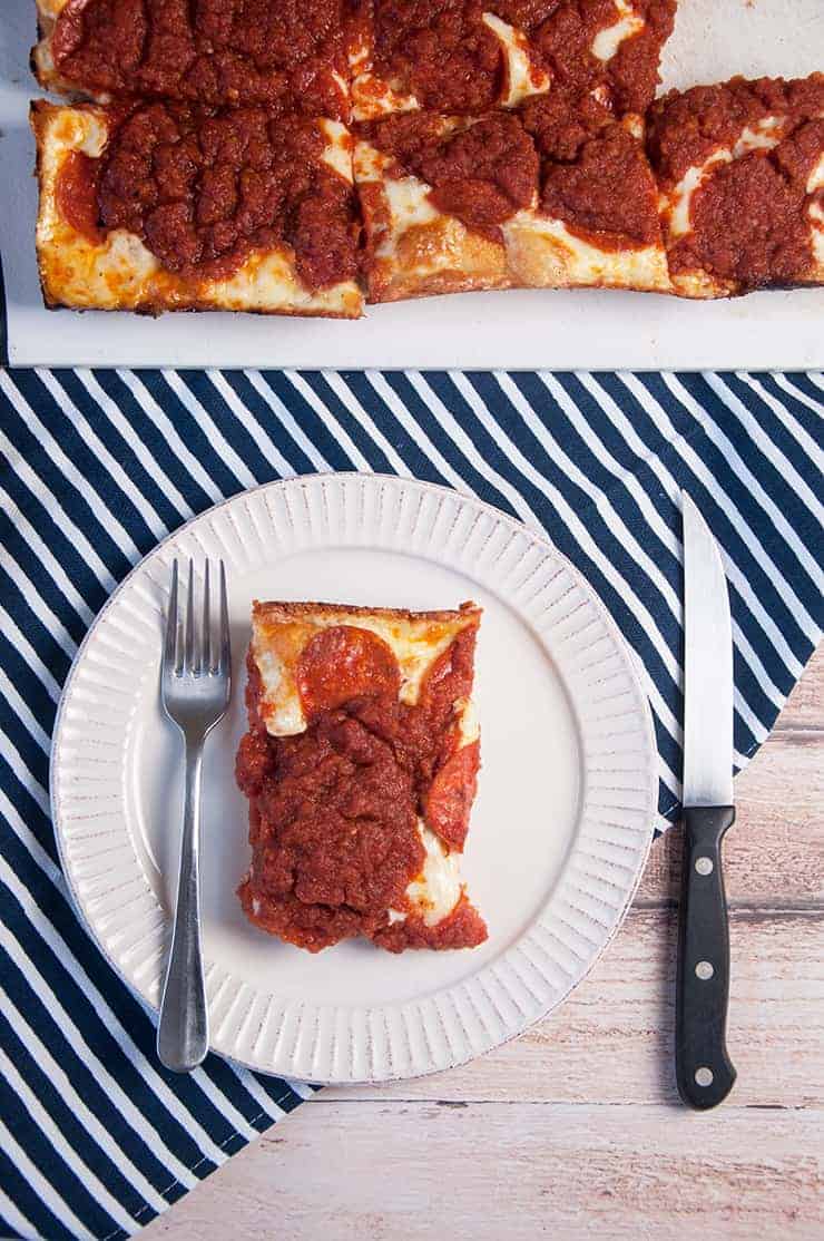 One Slice of Detroit Style Pizze on a white plate with the full pizza at the top on a cutting board.